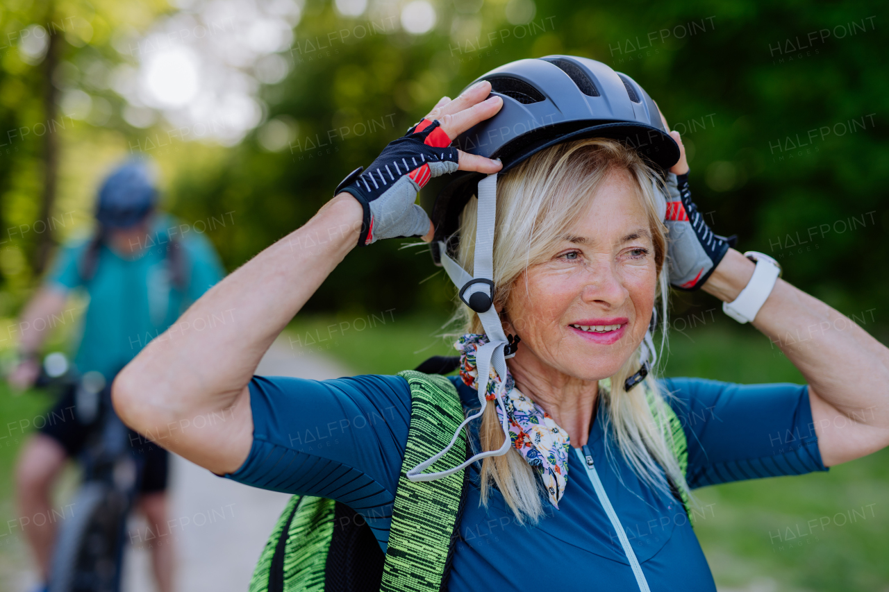 An active senior couple riding bicycles at summer park, woman is putting on helmet, healthy lifestyle concept.