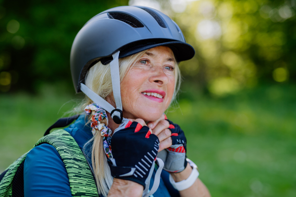 An active senior couple riding bicycles at summer park, woman is putting on helmet, healthy lifestyle concept.