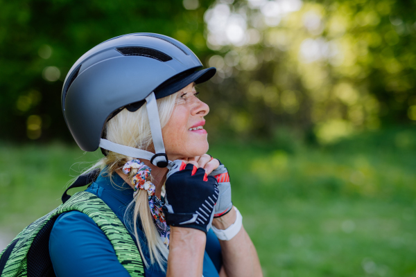 An active senior couple riding bicycles at summer park, woman is putting on helmet, healthy lifestyle concept.