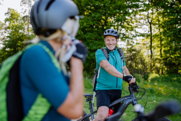 An active senior couple riding bicycles at summer park, woman is putting on helmet, healthy lifestyle concept.