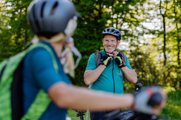 aN Active senior couple riding bicycles at summer park, man is putting on helmet, healthy lifestyle concept.