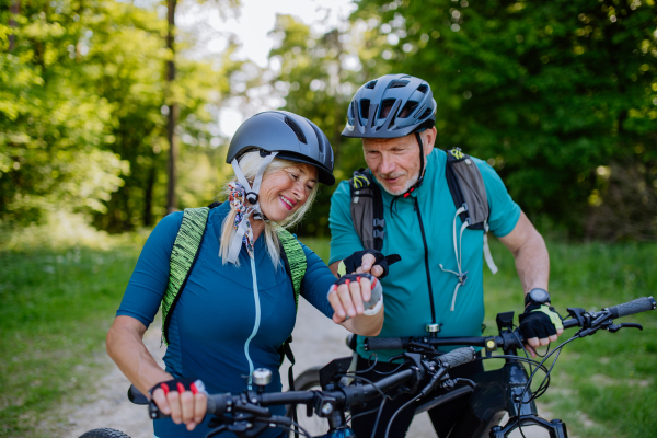 An active senior couple riding electric bicycles on road at summer park, checking their performance at smartwatch, healthy lifestyle concept.