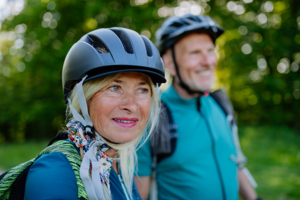 An active senior couple riding bicycles at summer park, woman with bicycle helmet, healthy lifestyle concept.