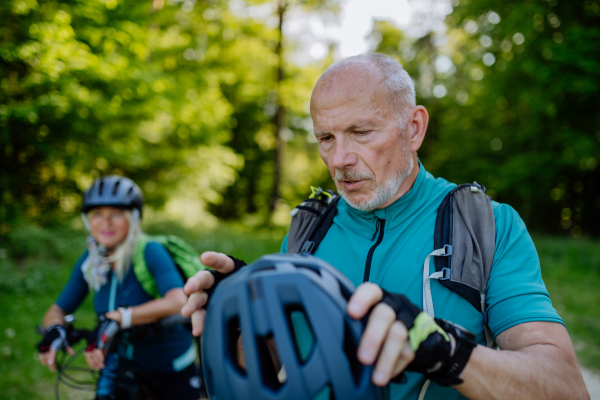 An active senior couple riding bicycles at summer park, man is putting on helmet, healthy lifestyle concept.