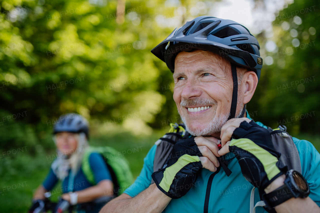 aN Active senior couple riding bicycles at summer park, man is putting on helmet, healthy lifestyle concept.