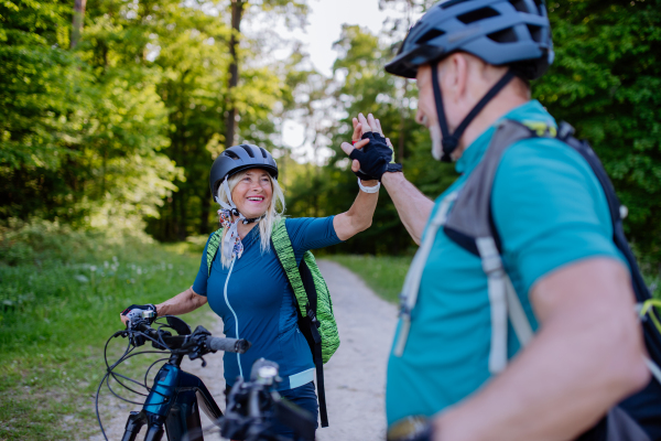 An active senior couple riding electric bicycles on road at summer park, high fiving, healthy lifestyle concept.