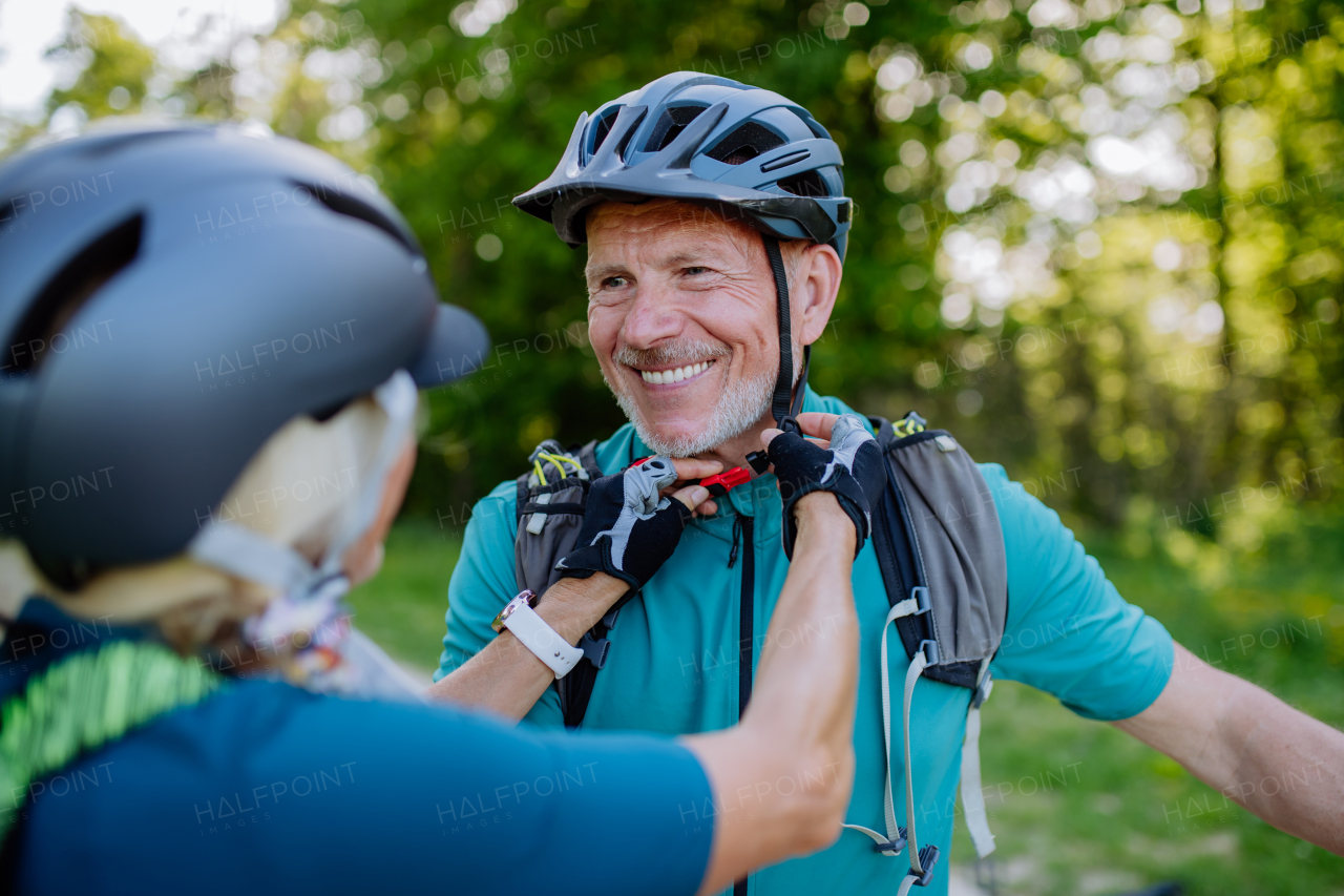 aN Active senior couple riding bicycles at summer park, man is putting on helmet, healthy lifestyle concept.