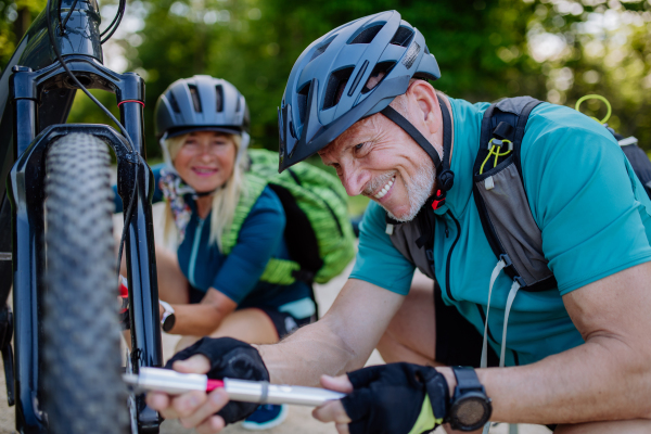 An active senior couple repairing bicycle, pumping up tire in nature in summer.