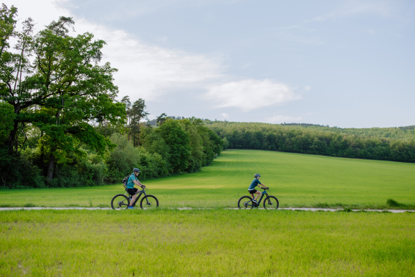 An active senior couple riding electric bicycles on trail at summer park, healthy lifestyle concept. Side view.