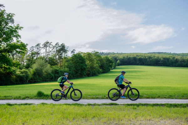 An active senior couple riding electric bicycles on path at summer park, healthy lifestyle concept. Side view.