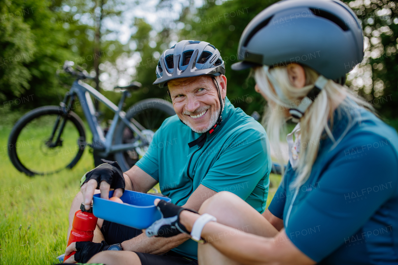 An active senior couple resting after bicycle ride at summer park, sitting on grass and having snack.