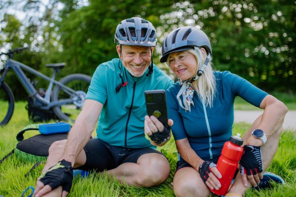 An active senior couple resting after bicycle ride at summer park, sitting on grass and using smartphone.