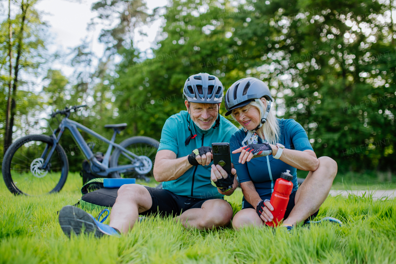 An active senior couple resting after bicycle ride at summer park, sitting on grass and using smartphone.