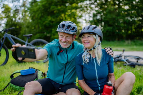 An active senior couple resting after bicycle ride at summer park, sitting on grass and taking slefie.