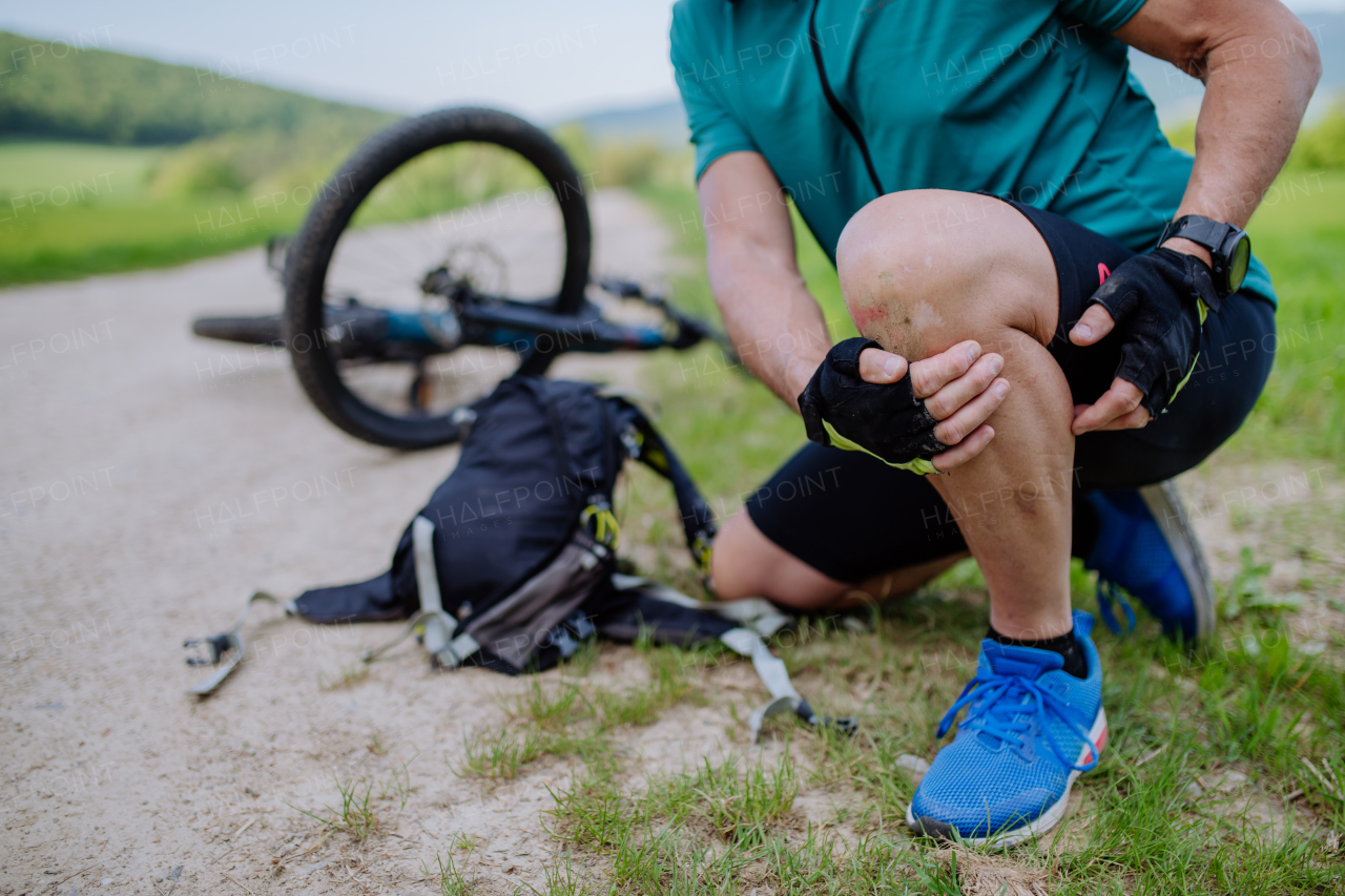 An active senior man in sportswear fell off bicycle on the ground and hurt his knee, in park in summer.