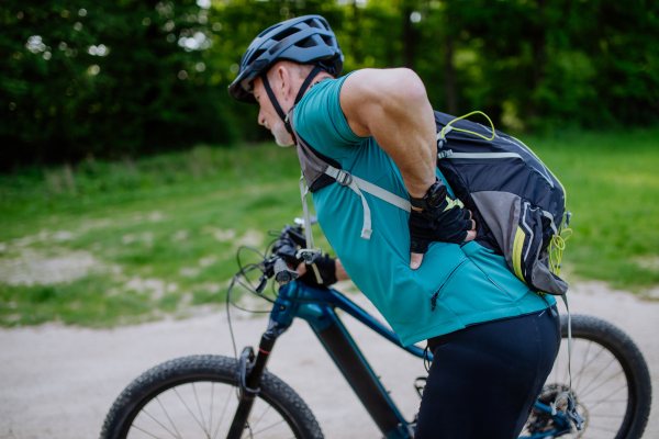 An active senior man in sportswear suffiering from pain in his back after cycling, in park in summer.