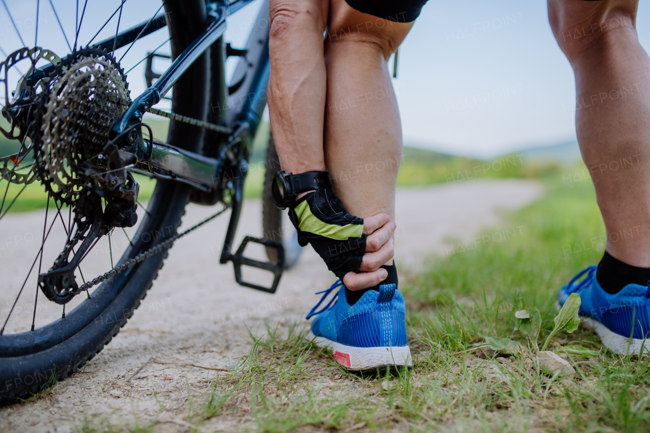 A close-up of active senior man in sportswear suffiering from pain in his ankle after cycling, in park in summer.