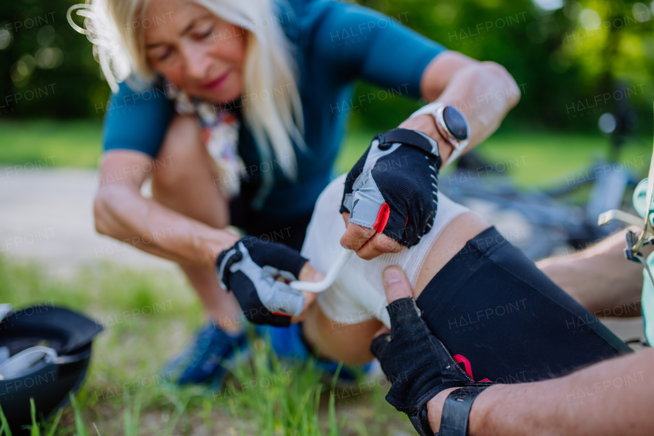 A senior woman is helping man after he fell off bicycle on the ground and injured his knee, in park in summer.