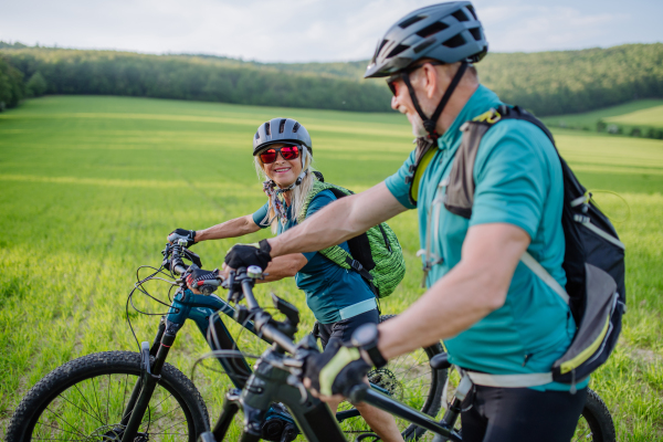 An active senior couple with electric bicycles on meadow in summer, healthy lifestyle concept.