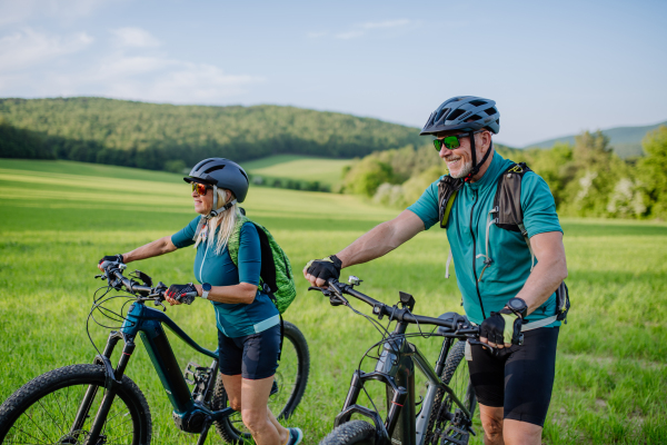 An active senior couple pushing electric bicycles on grass trail at summer park, healthy lifestyle concept.