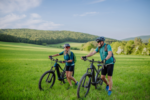 An active senior couple with electric bicycles on meadow in summer, healthy lifestyle concept.