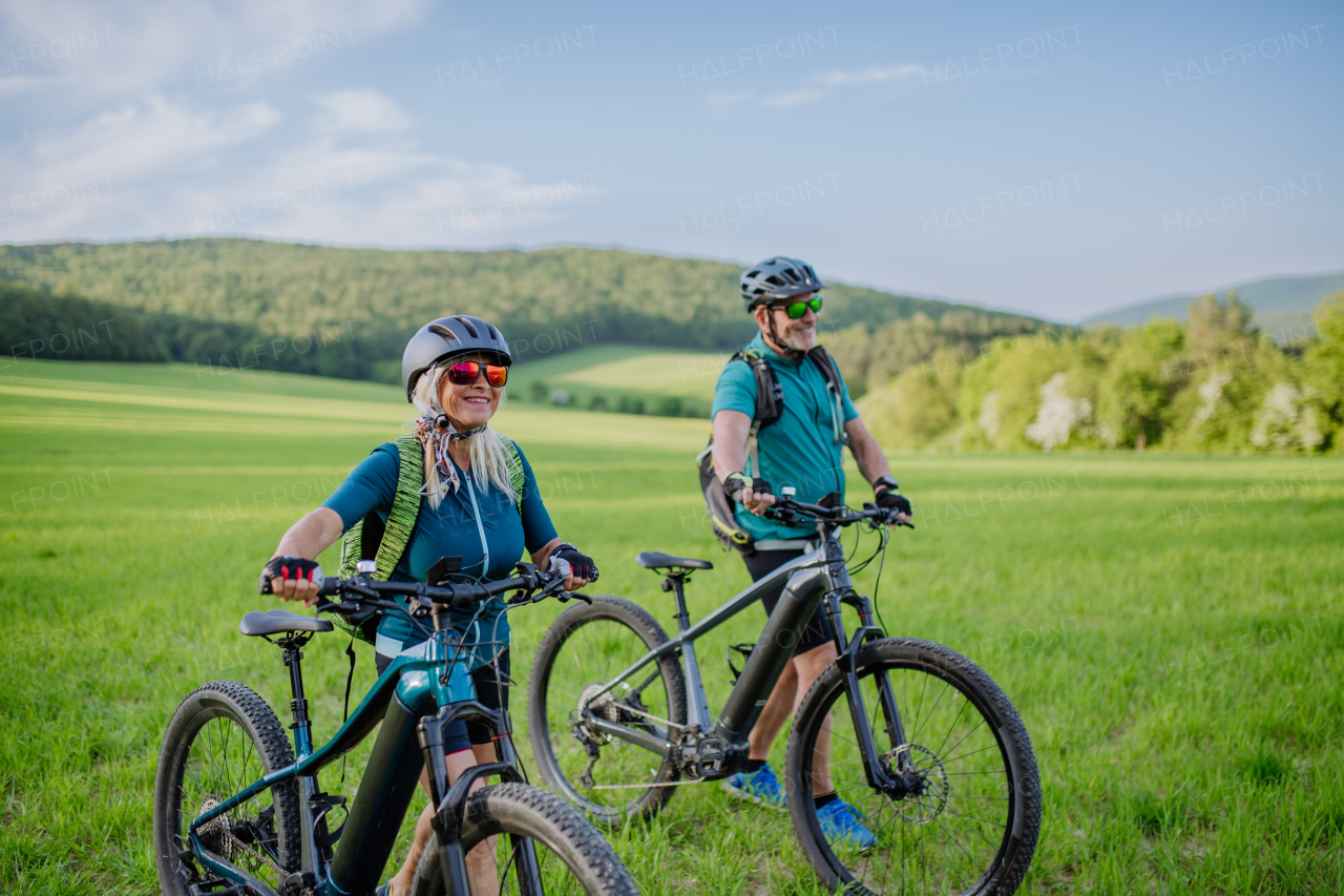 An active senior couple with electric bicycles on meadow in summer, healthy lifestyle concept.