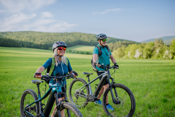 An active senior couple with electric bicycles on meadow in summer, healthy lifestyle concept.