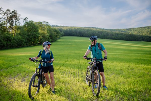 An active senior couple with electric bicycles on meadow in summer, healthy lifestyle concept.