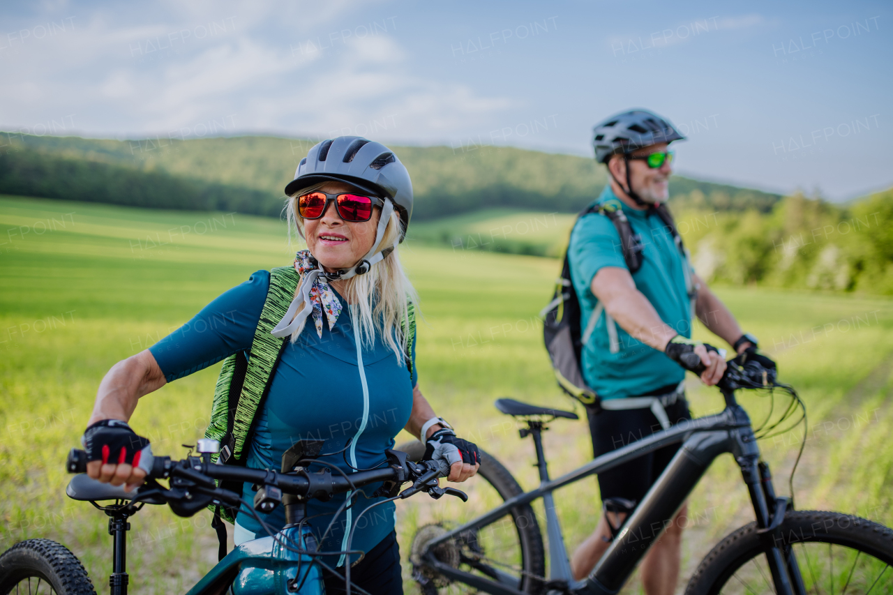 An active senior couple with electric bicycles on meadow in summer, healthy lifestyle concept.