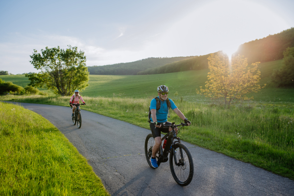 An active senior couple riding electric bicycles on path at summer park, healthy lifestyle concept. Rear view.