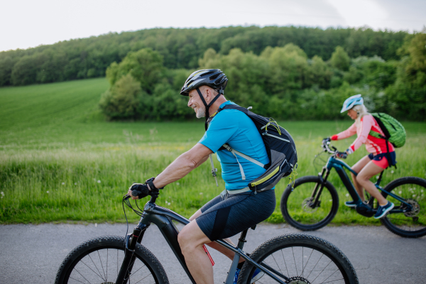 An active senior couple riding electric bicycles on path at summer park, healthy lifestyle concept. Side view.