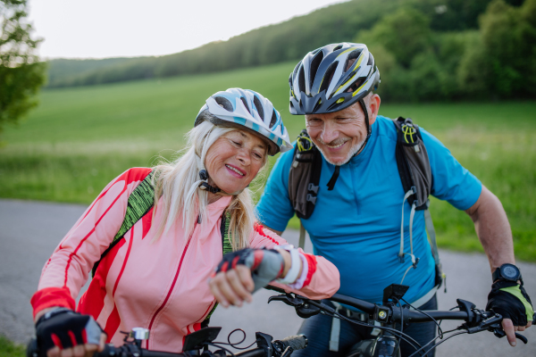 An active senior couple riding electric bicycles on road at summer park, checking their performance at smartwatch, healthy lifestyle concept.