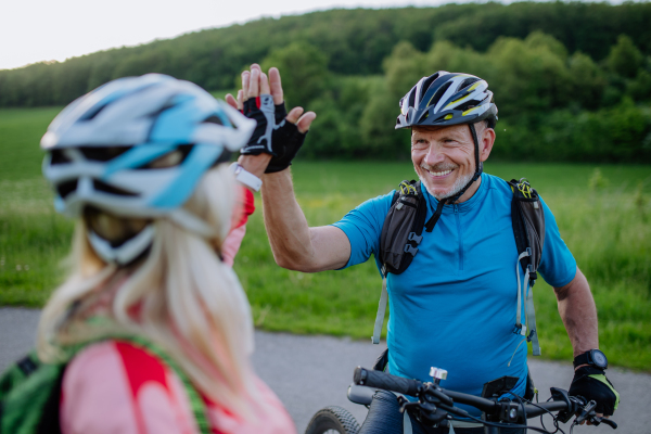 An active senior couple riding electric bicycles on road at summer park, high fiving, healthy lifestyle concept.