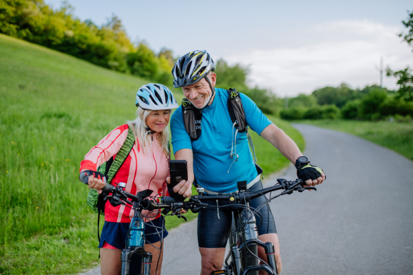 An active senior couple resting after bicycle ride at summer park, using smartphone.