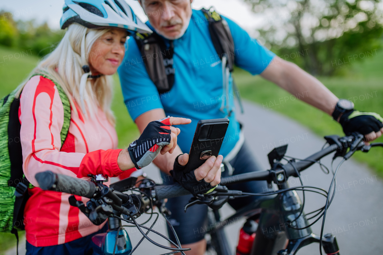 An active senior couple resting after bicycle ride at summer park, using smartphone.