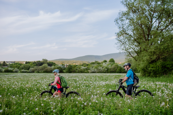 An active senior couple with electric bicycles on meadow in summer, healthy lifestyle concept.