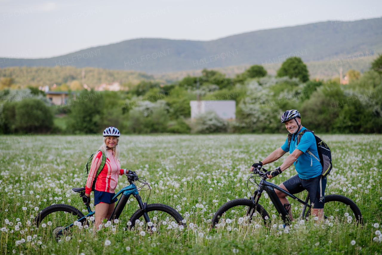 An active senior couple with electric bicycles on meadow in summer, healthy lifestyle concept.