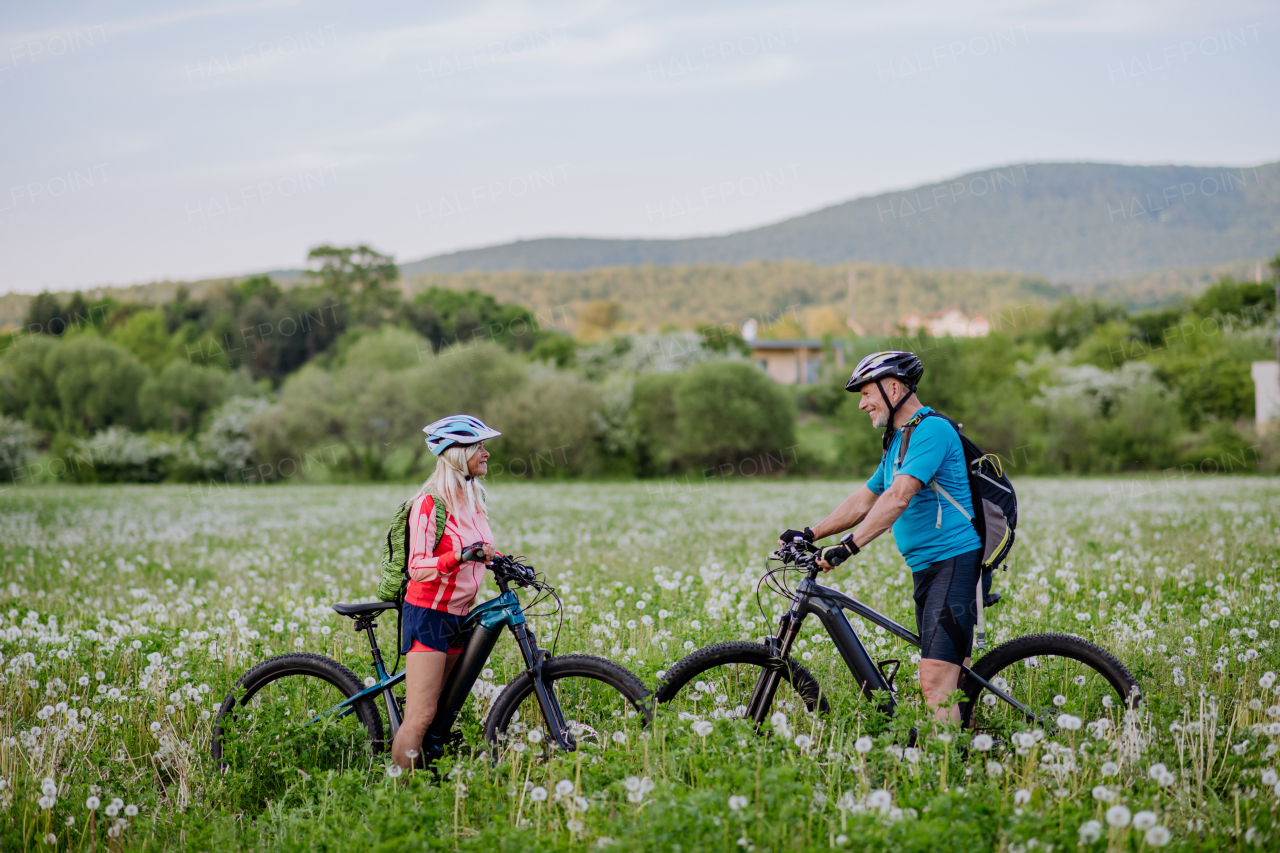 An active senior couple with electric bicycles on meadow in summer, healthy lifestyle concept.