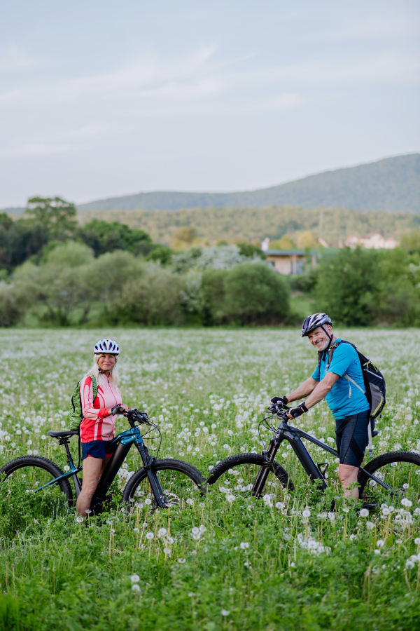 An active senior couple with electric bicycles on meadow in summer, healthy lifestyle concept.