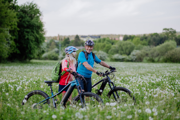 An active senior couple with electric bicycles on meadow in summer, healthy lifestyle concept.