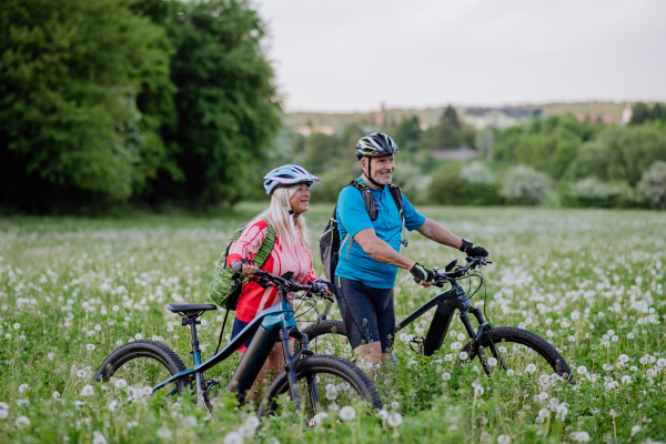 An active senior couple with electric bicycles on meadow in summer, healthy lifestyle concept.