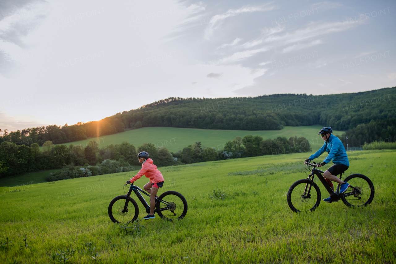 An active senior couple riding electric bicycles on trail at summer park, healthy lifestyle concept. Side view.