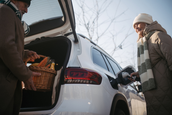 Senior woman charging their electric car, during his husban giving purchase in a car trunk.