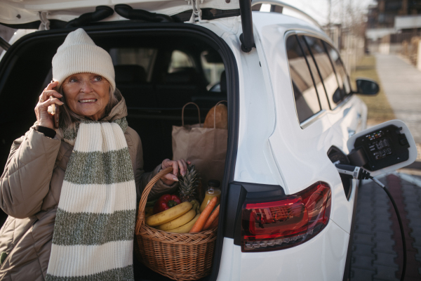 Senior woman sitting in car trunk with grocery purchase and waiting for charging her electric car.