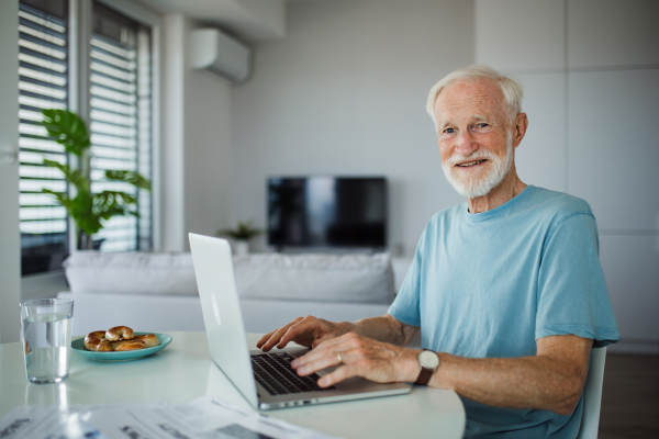 Senior man in a wheelchair working from home during retirement. Elderly man using digital technologies, working on a laptop. Concept of seniors and digital skills.