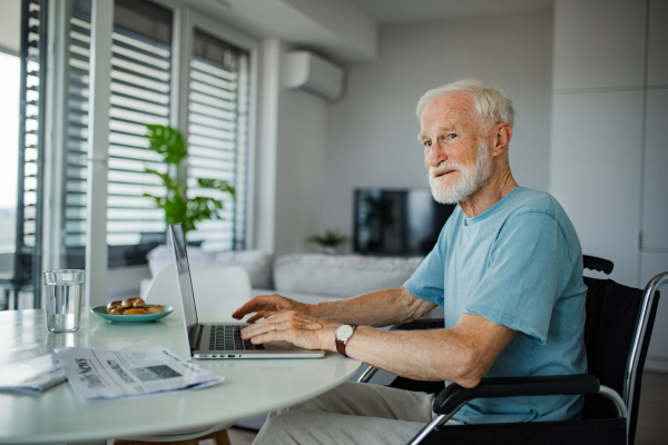 Senior man in a wheelchair shopping online on laptop. Elderly man using digital technologies, working on a notebook in his kitchen. Concept of seniors and digital skills.