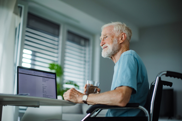 Senior man in a wheelchair working from home during retirement. Elderly man using digital technologies, working on a laptop, videocalling someone. Concept of seniors and digital skills.