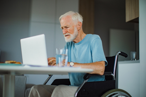 Senior man in a wheelchair working from home during retirement. Elderly man using digital technologies, working on a laptop. Concept of seniors and digital skills.