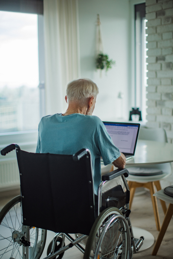 Senior man in a wheelchair writting document on laptop. Elderly man using digital technologies, working on a notebook in his kitchen. Concept of seniors and digital skills.