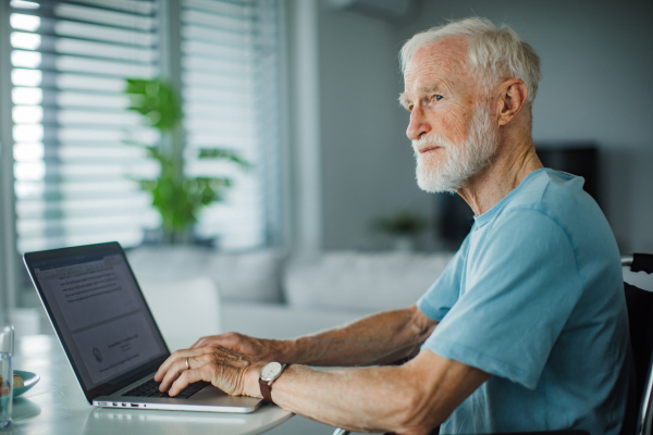 Senior man in a wheelchair working from home during retirement. Portrait of elderly man using digital technologies, working on a laptop. Concept of seniors and digital skills.